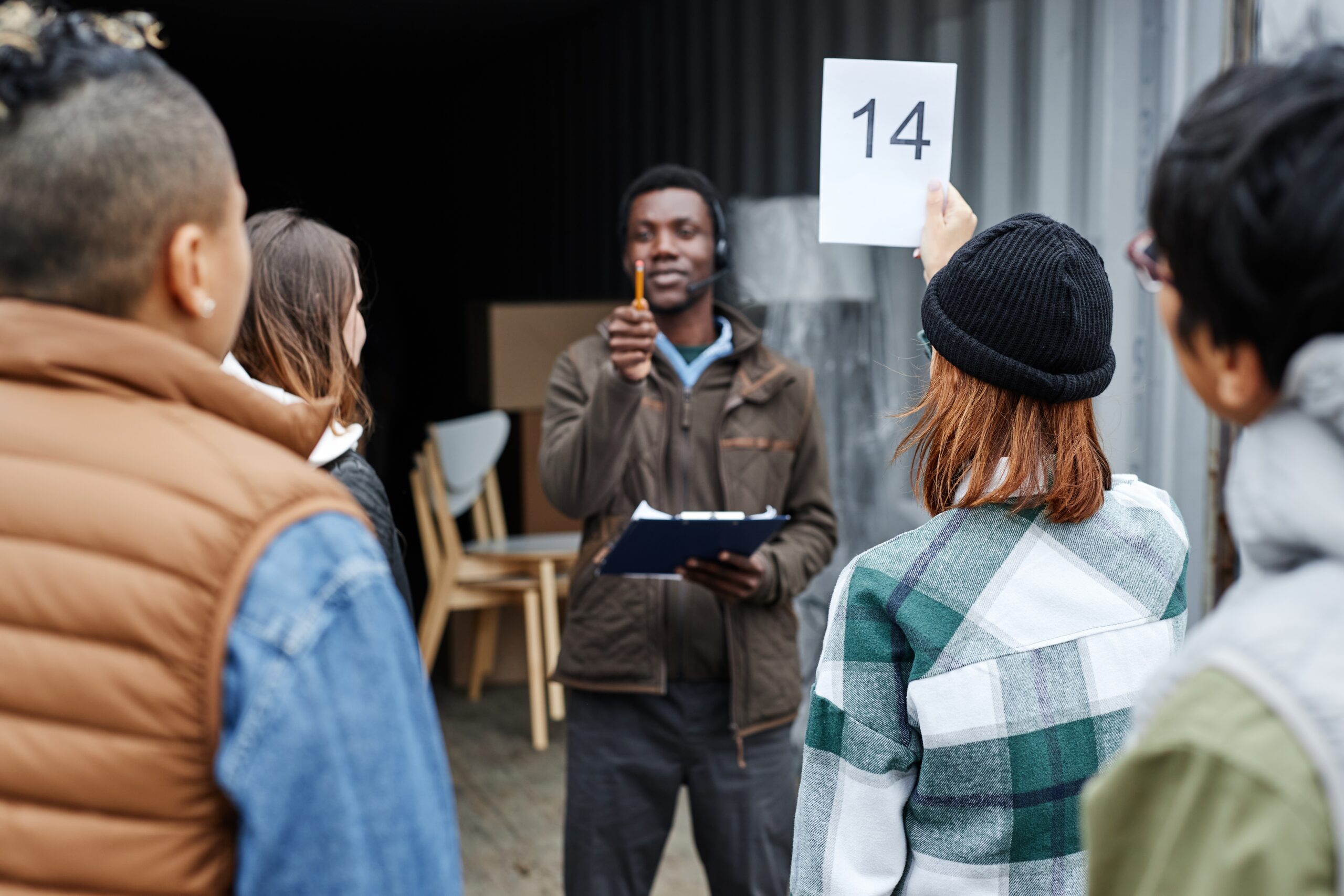 An auctioneer holding a clipboard guides a crowd of buyers outside an open storage locker as one participant raises a bidding card numbered 14. This live, on-site environment demonstrates the traditional, physical process of how do storage auctions work for unpaid units.